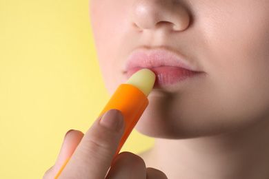 Photo of Woman applying hygienic lipstick on lips against yellow background, closeup