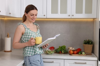 Photo of Happy woman with recipe book and cup of tea in kitchen, space for text