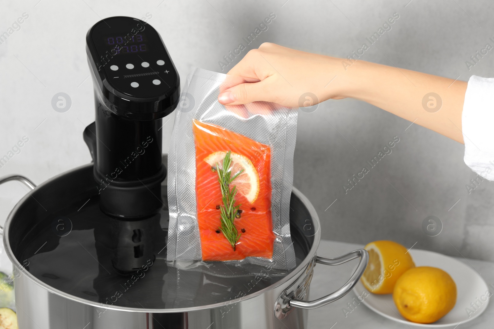 Photo of Woman putting vacuum packed salmon into pot with sous vide cooker in kitchen, closeup. Thermal immersion circulator