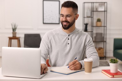 Photo of Young man writing in notebook while working on laptop at white table indoors