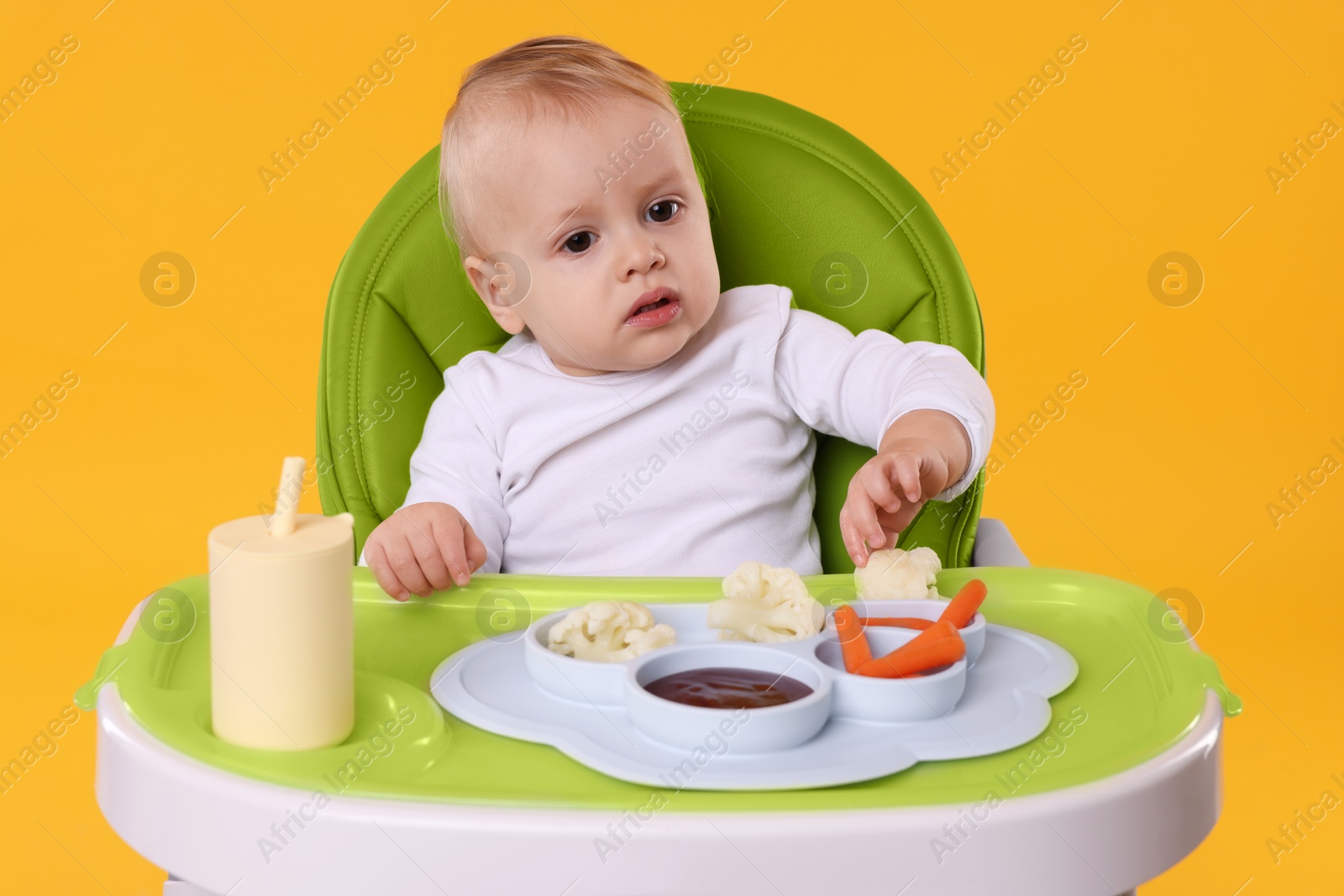 Photo of Cute little baby with healthy food in high chair on orange background