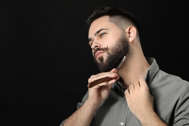 Handsome young man shaving beard with blade on black background. Space for text