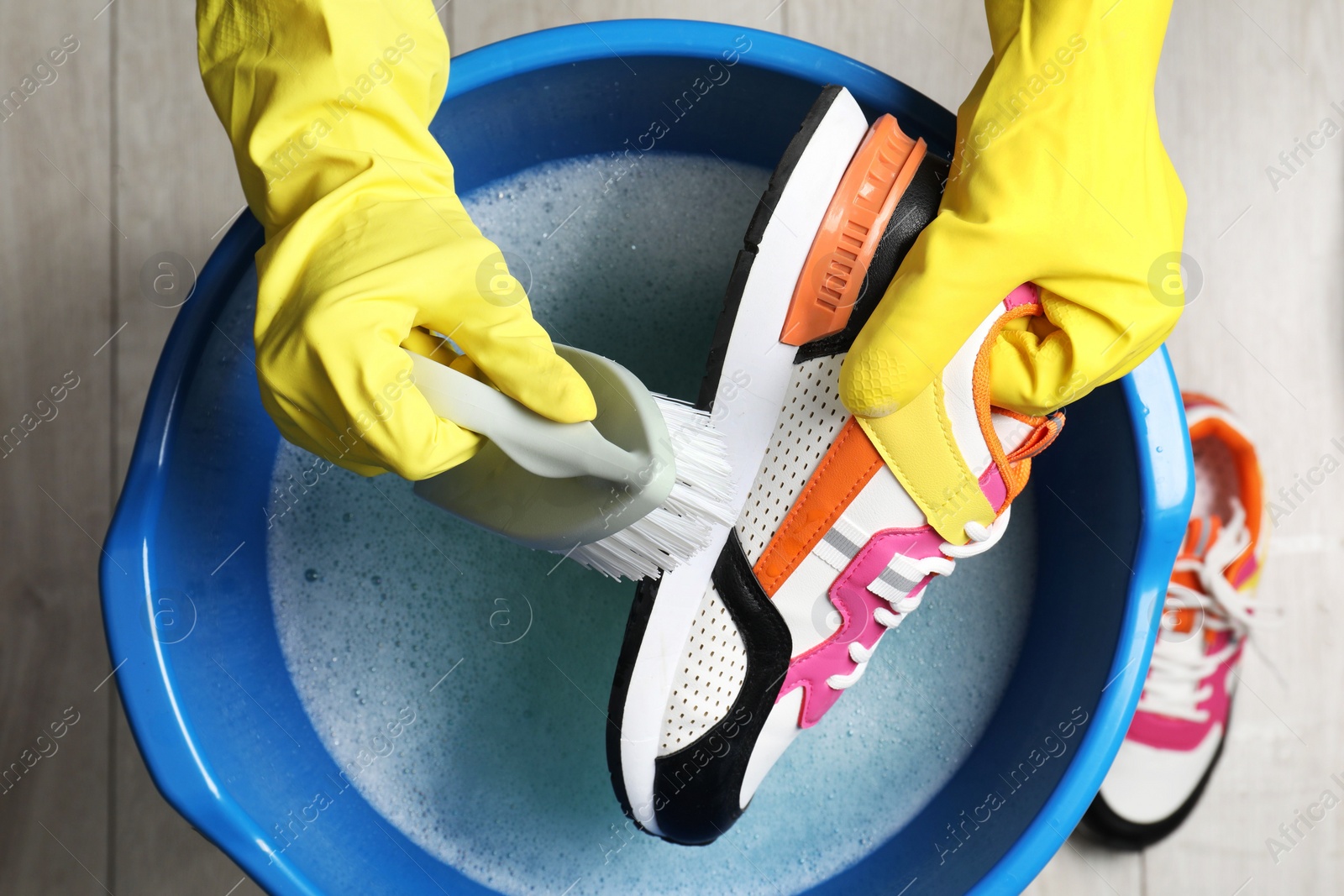 Photo of Woman with gloves and brush cleaning stylish sneakers in wash basin, top view