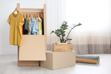 Cardboard wardrobe box with clothes on hangers, houseplant and carpet indoors. Moving day