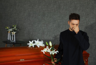 Sad young man with white lilies near casket in funeral home
