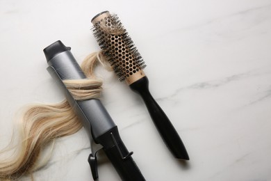 Curling iron and round brush with blonde hair lock on white marble background, flat lay. Space for text