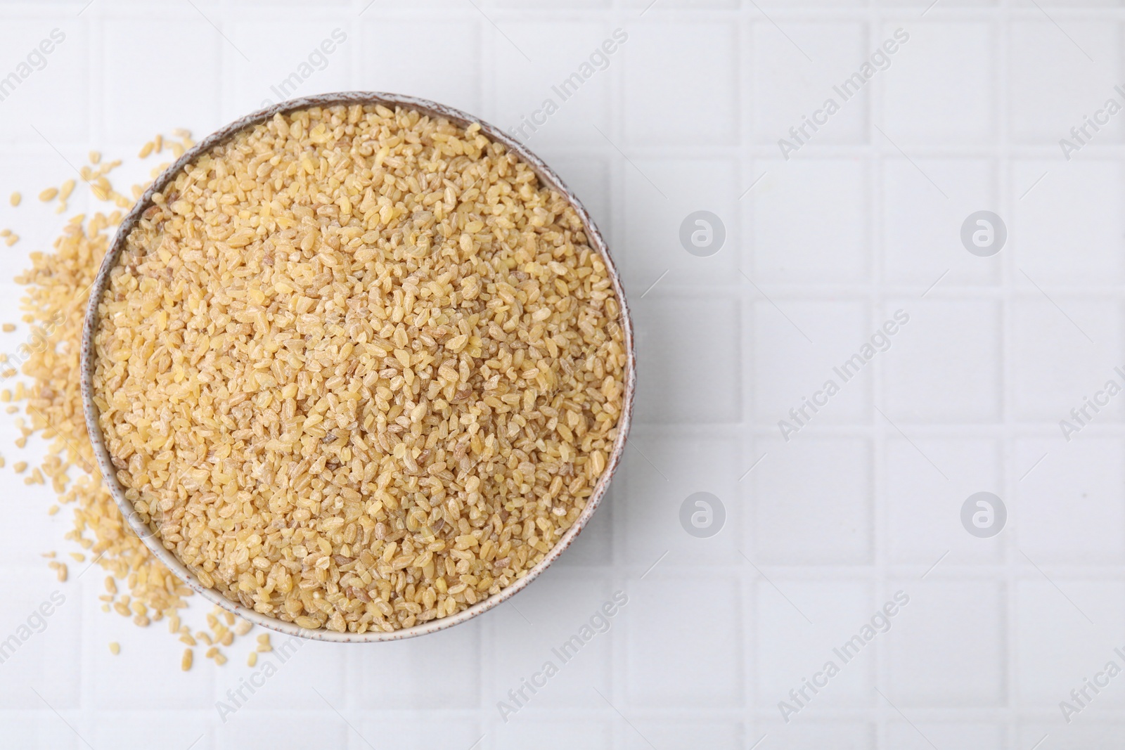 Photo of Raw bulgur in bowl on white tiled table, top view. Space for text