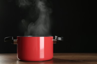 Photo of Pot with steam on wooden table against black background, space for text