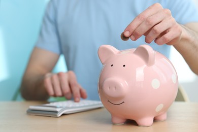 Financial savings. Man putting coin into piggy bank while using calculator at wooden table, closeup