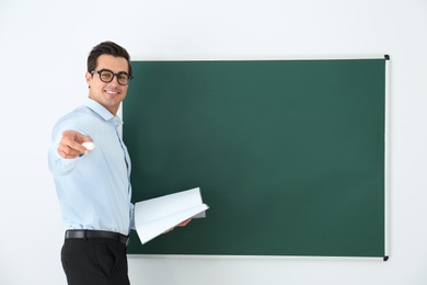Photo of Young teacher with book near blank chalkboard in classroom. Space for text