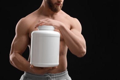 Young man with muscular body holding jar of protein powder on black background, closeup. Space for text