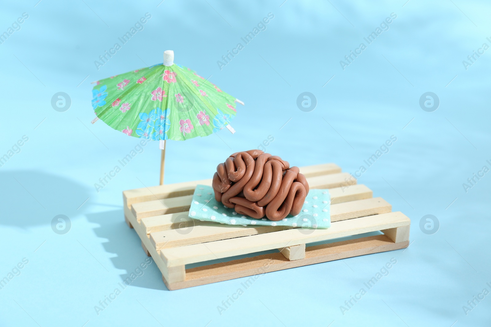 Photo of Brain made of plasticine on mini wooden sunbed under umbrella against light blue background