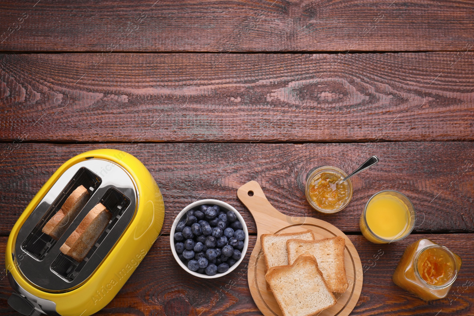 Photo of Yellow toaster with roasted bread, glass of juice, blueberries and jam on wooden table, flat lay. Space for text