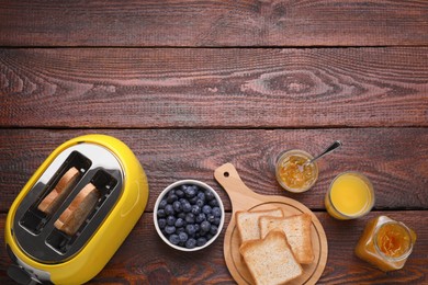 Photo of Yellow toaster with roasted bread, glass of juice, blueberries and jam on wooden table, flat lay. Space for text