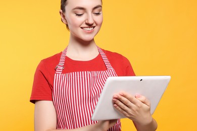 Photo of Beautiful young woman in clean striped apron with tablet on orange background, closeup