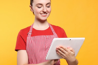 Beautiful young woman in clean striped apron with tablet on orange background, closeup