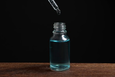 Photo of Dripping cosmetic serum from pipette into bottle at wooden table against black background