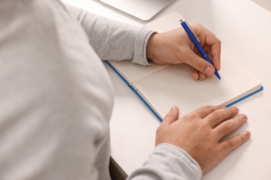 Young man writing in notebook at white table, closeup