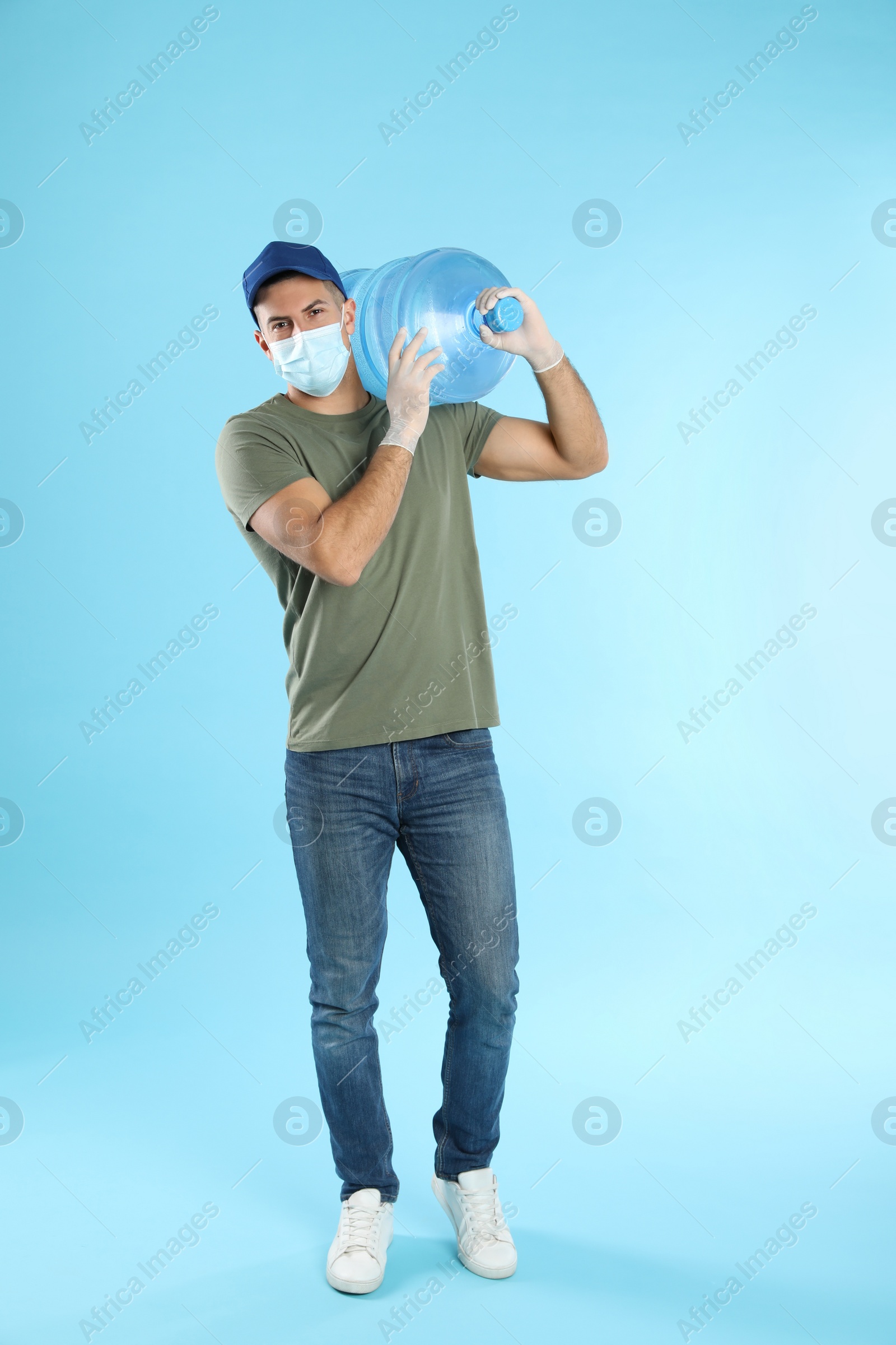 Photo of Courier in face mask with bottle of cooler water on light blue background. Delivery during coronavirus quarantine