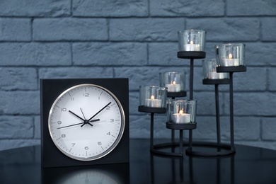 Stylish analog clock and candles on table near brick wall