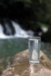 Photo of Glass of water on stone near waterfall outdoors. Space for text