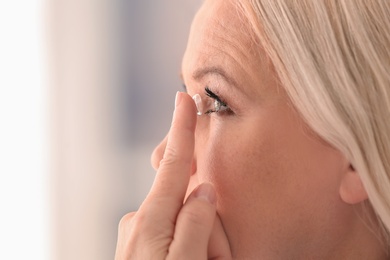 Senior woman putting contact lens in her eye on blurred background