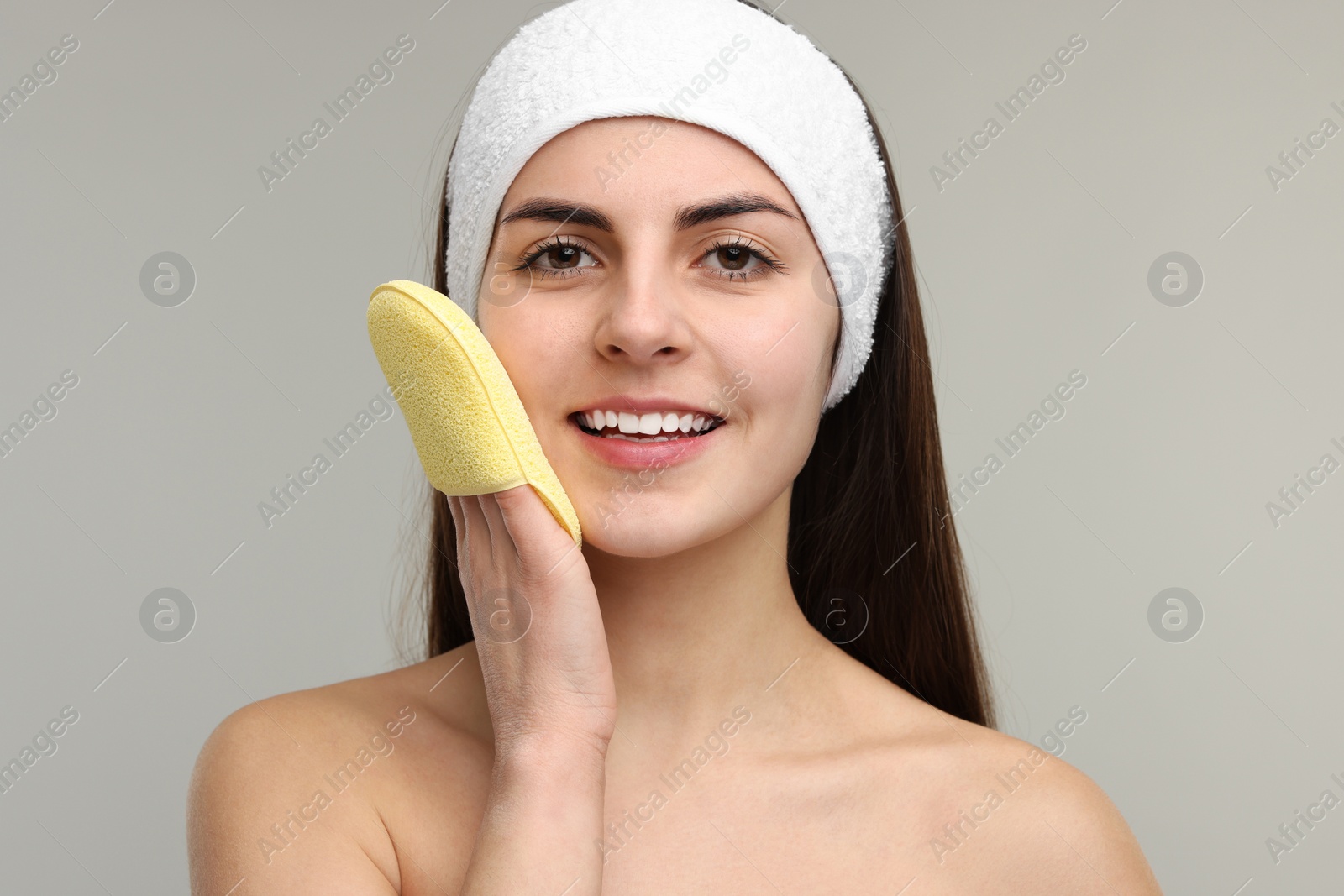 Photo of Young woman with headband washing her face using sponge on light grey background