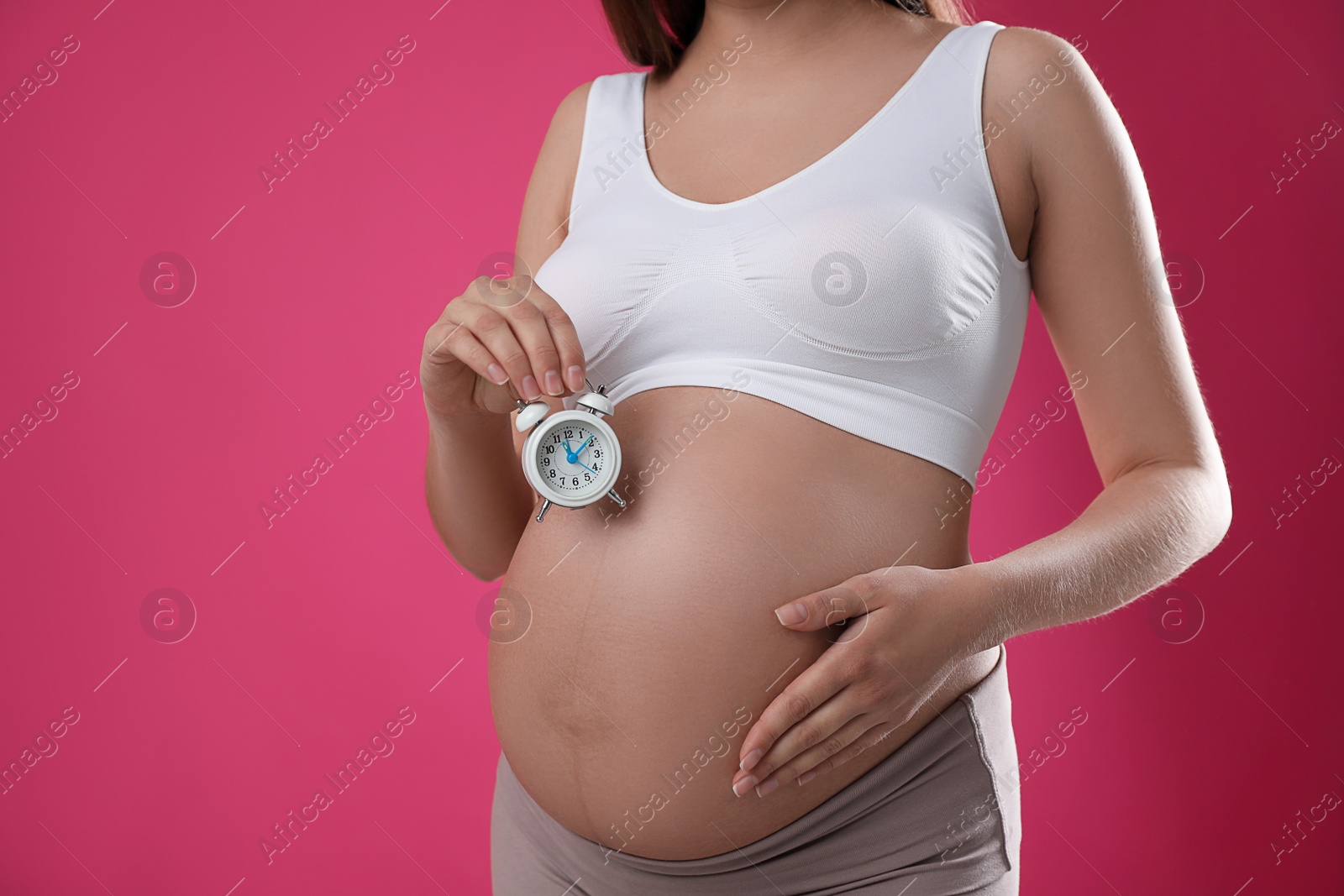 Photo of Young pregnant woman holding alarm clock near her belly on pink background, closeup. Time to give birth