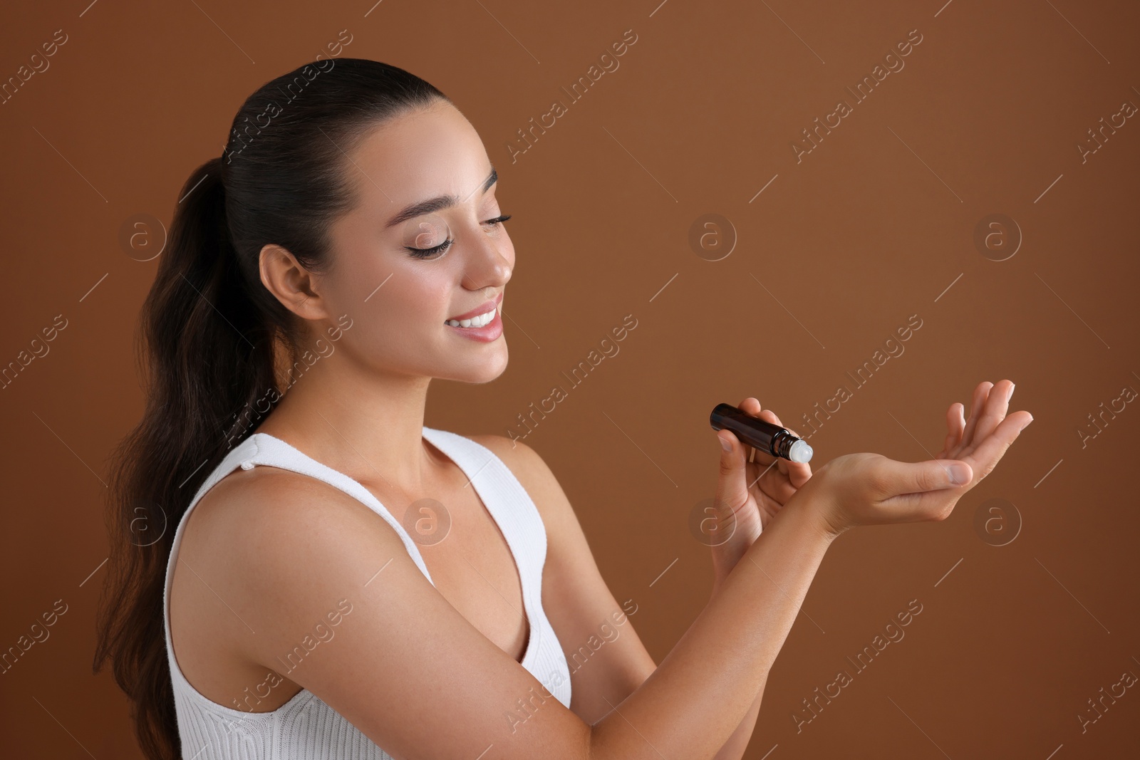 Photo of Beautiful happy woman with roller bottle applying essential oil onto wrist on brown background