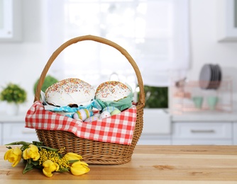 Image of Basket with delicious Easter cakes, painted eggs and flowers on wooden table indoors. Space for text