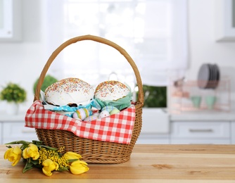 Basket with delicious Easter cakes, painted eggs and flowers on wooden table indoors. Space for text