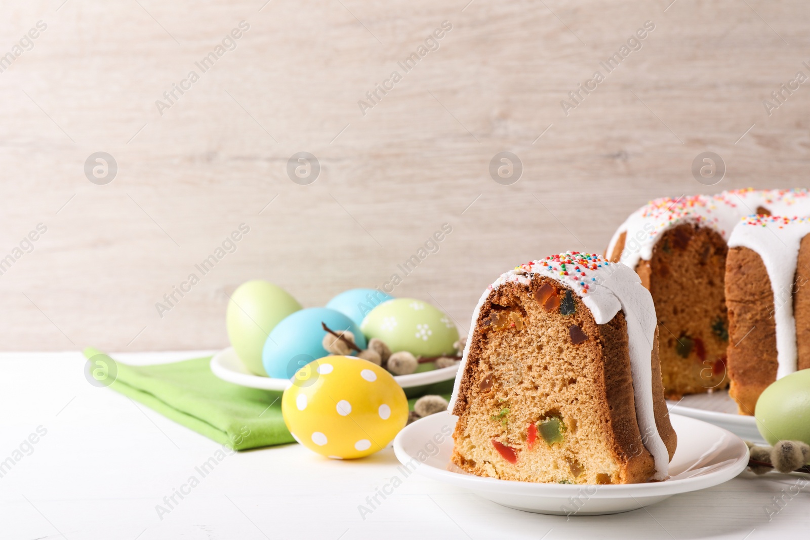 Photo of Piece of glazed Easter cake with sprinkles and painted eggs on white wooden table, space for text