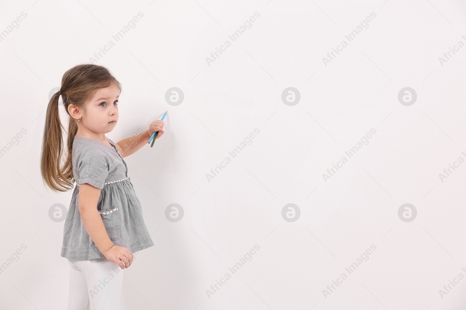Photo of Little girl drawing with colorful pencils on white wall indoors. Space for text