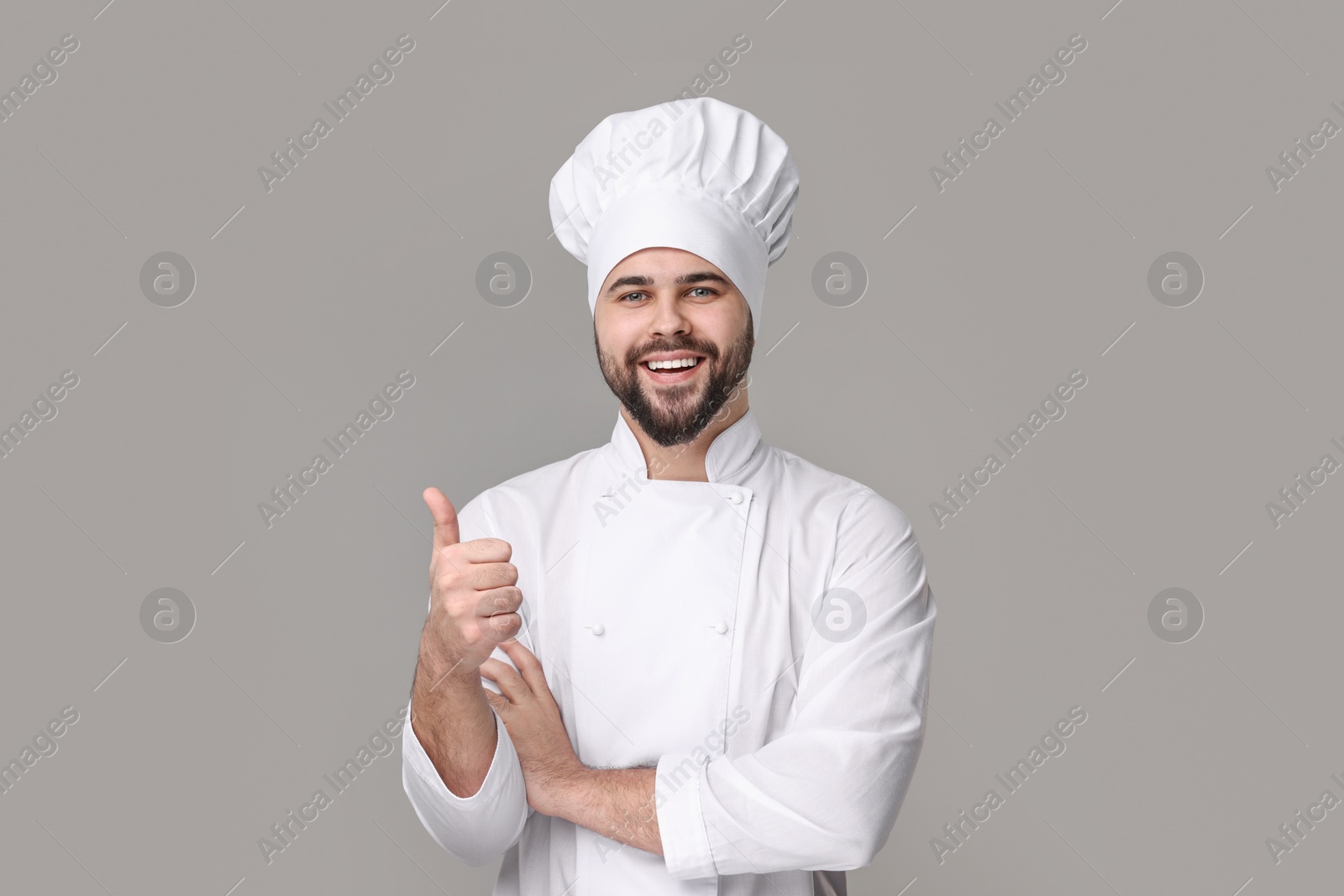 Photo of Happy young chef in uniform showing thumb up on grey background