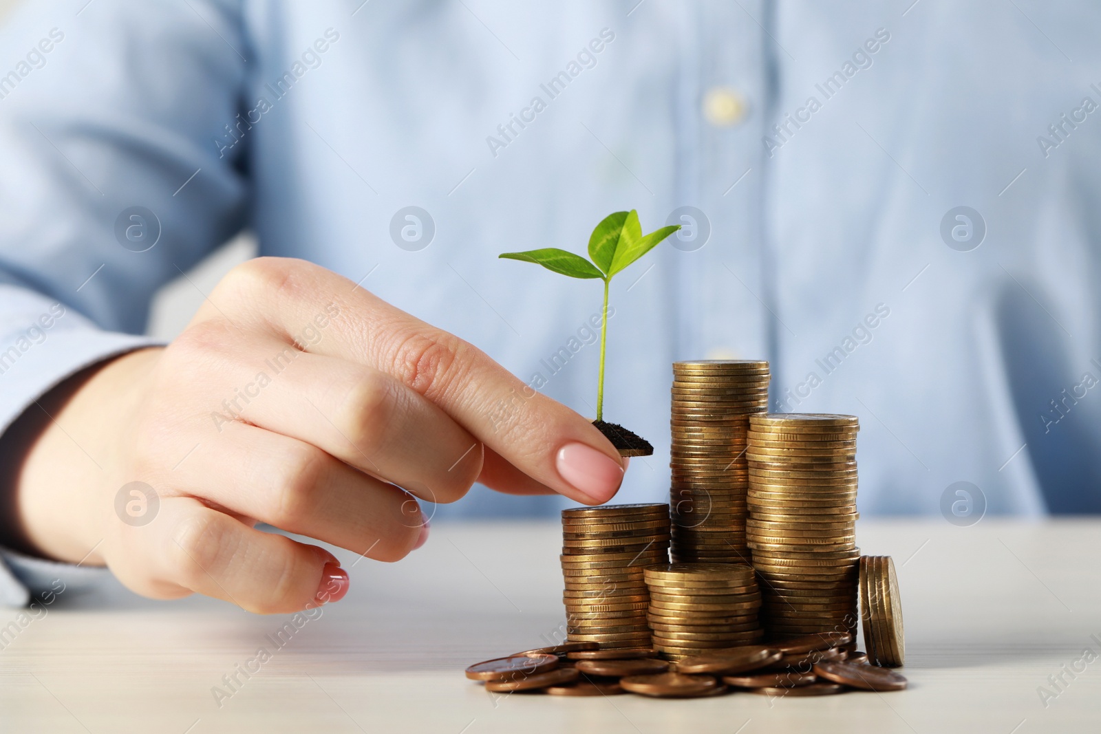 Photo of Woman putting coin with green sprout onto stack at white table, closeup. Investment concept