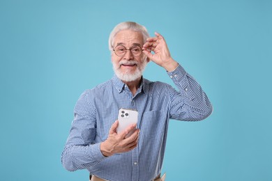 Photo of Portrait of stylish grandpa with glasses using smartphone on light blue background