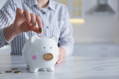 Photo of Woman putting money into piggy bank at white marble table indoors, closeup. Space for text