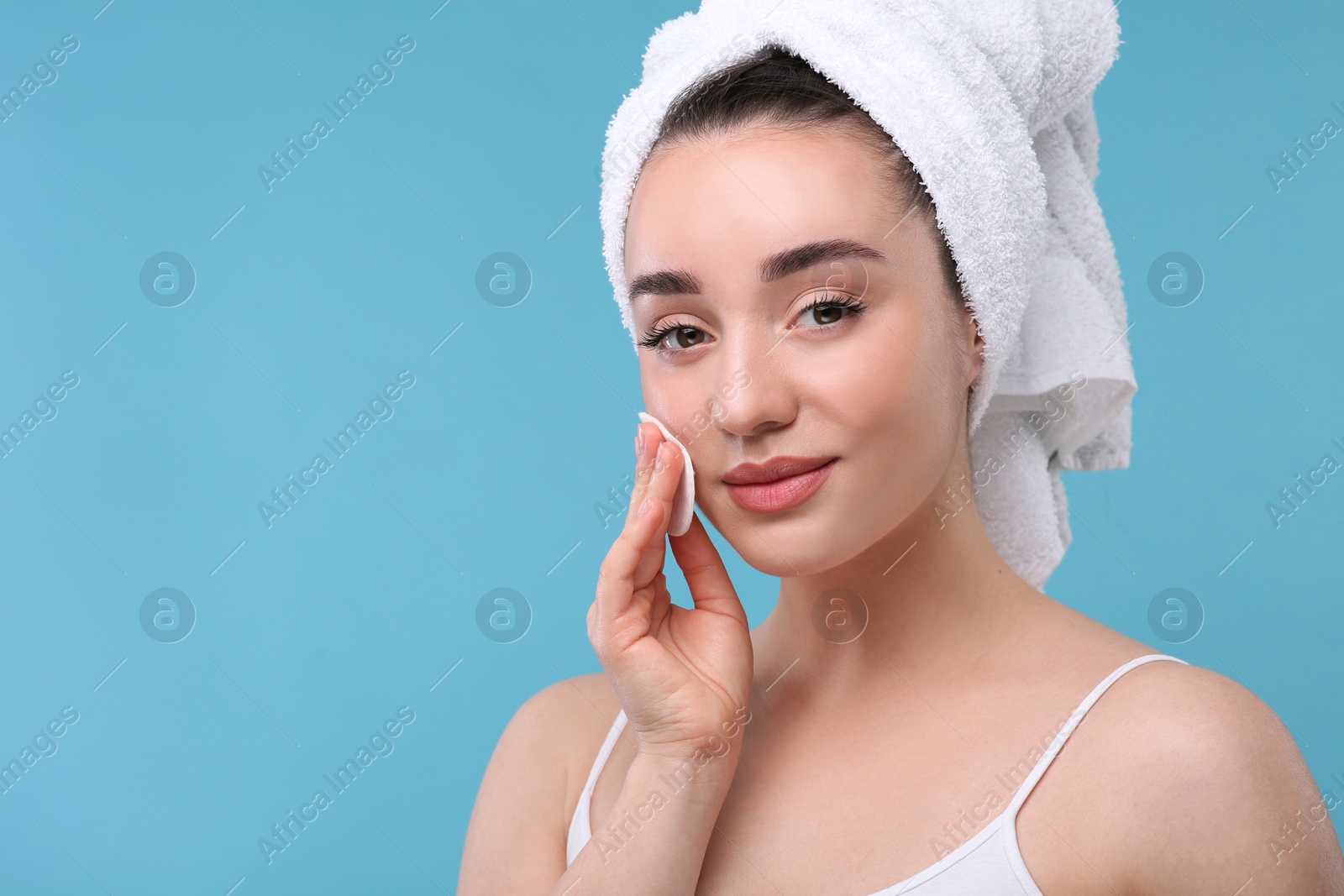 Photo of Beautiful woman in terry towel removing makeup with cotton pad on light blue background, space for text