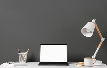 Photo of Stylish workplace with laptop, lamp and stationery on white table near grey wall