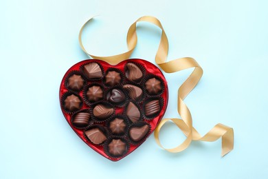 Heart shaped box with delicious chocolate candies and ribbon on light blue background, flat lay