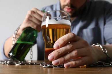 Addicted man with chained hands and alcoholic drink at wooden table, closeup