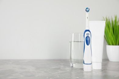 Electric toothbrush, tube with paste and glass of water on light grey marble table. Space for text