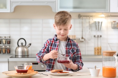 Photo of Cute little boy spreading jam onto tasty toasted bread at table in kitchen