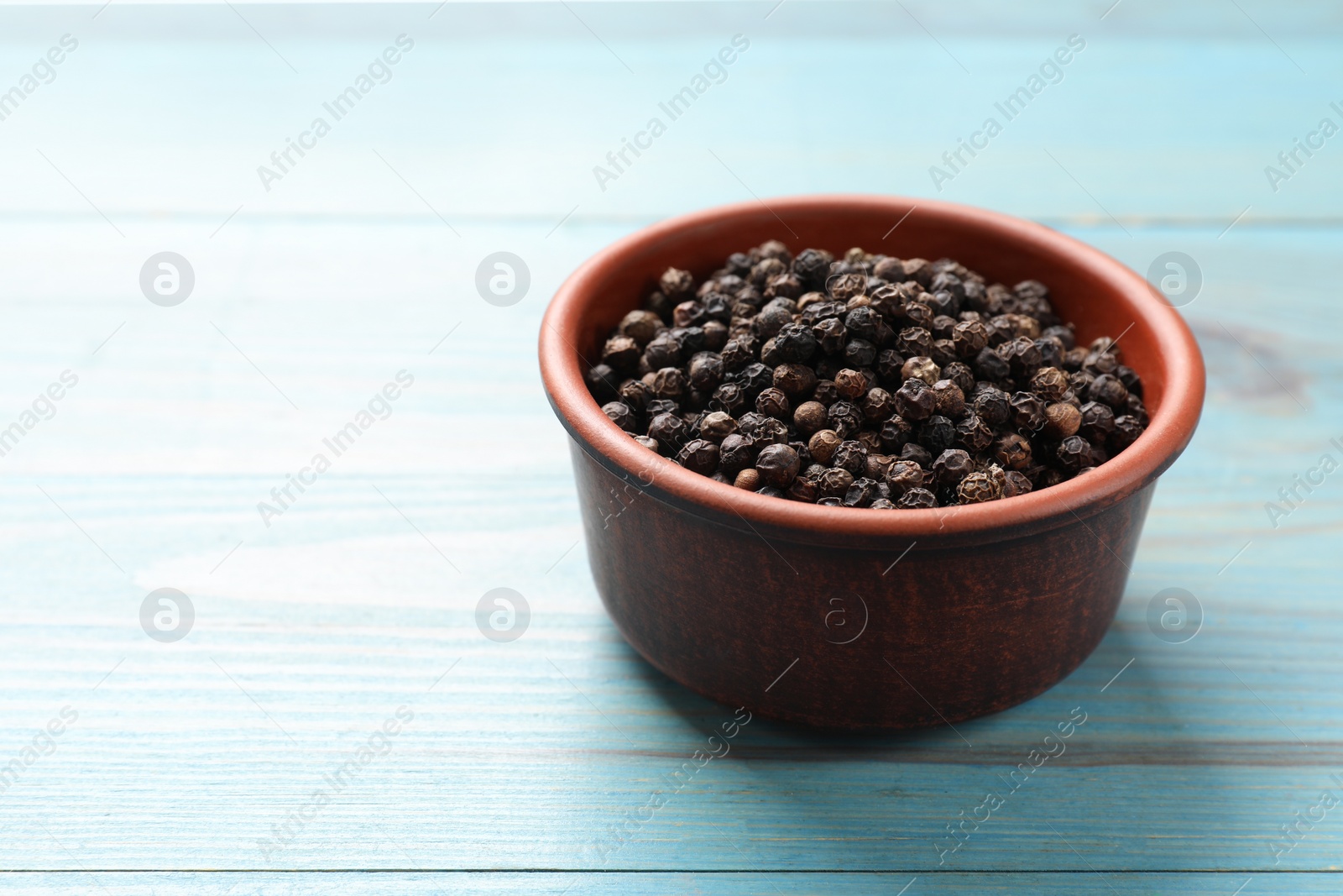 Photo of Aromatic spice. Black pepper in bowl on light blue wooden table, space for text