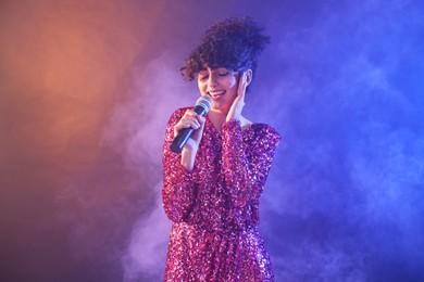 Beautiful young woman with microphone singing on color background in neon lights and smoke