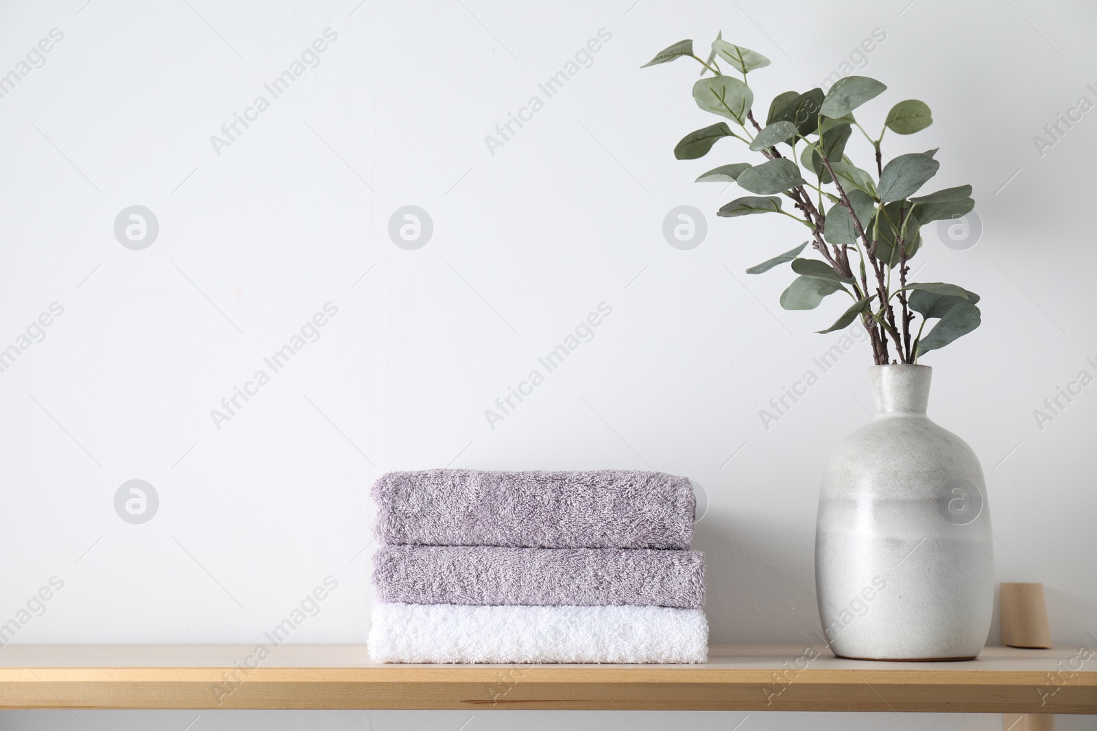 Photo of Stacked terry towels and eucalyptus branches in vase on wooden shelf near white wall, space for text