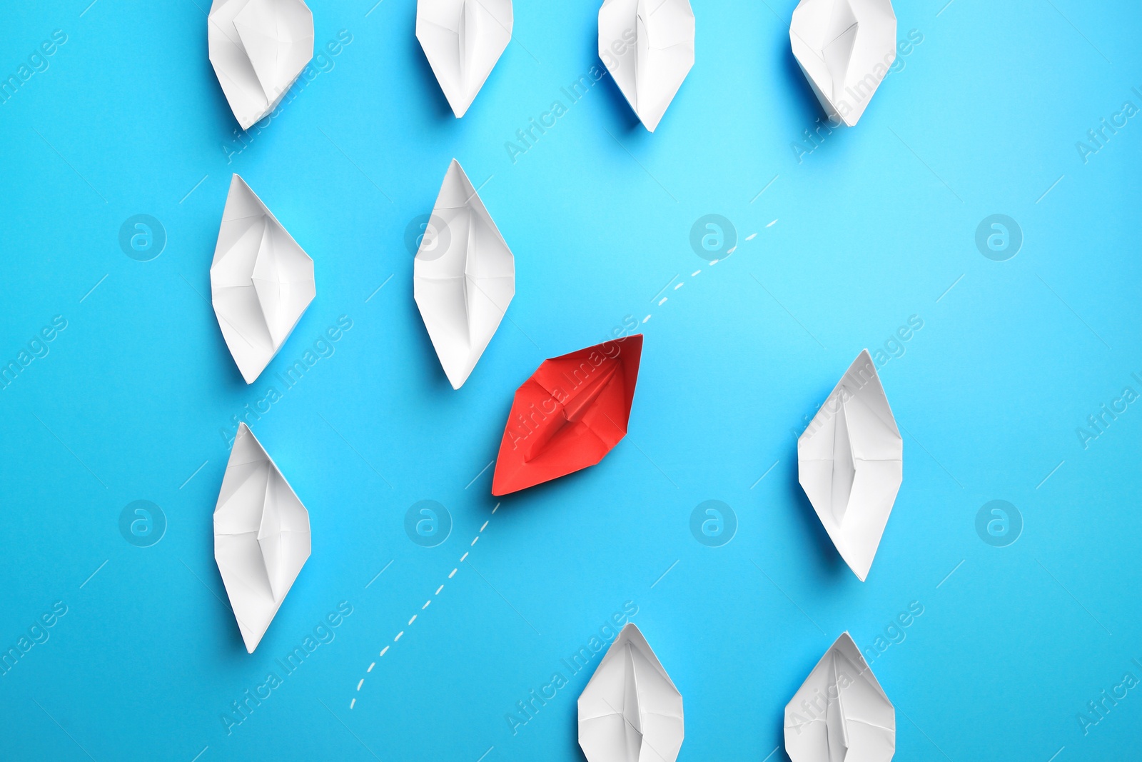 Photo of Red paper boat floating through others on light blue background, flat lay. Uniqueness concept