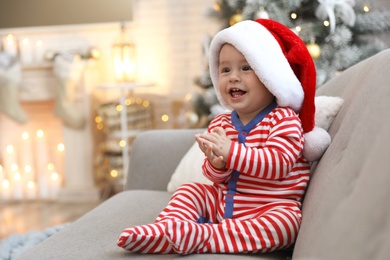 Cute baby in Santa hat sitting on sofa at home. Christmas celebration