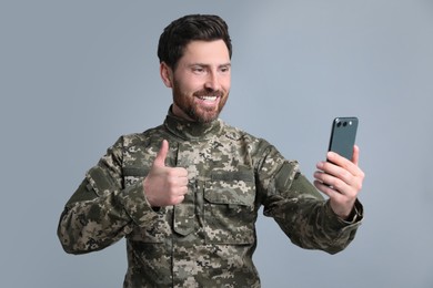 Photo of Happy soldier using video chat on smartphone and showing thumb up against light grey background. Military service