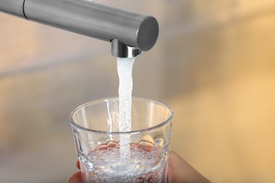 Photo of Woman filling glass with tap water from faucet on blurred background, closeup